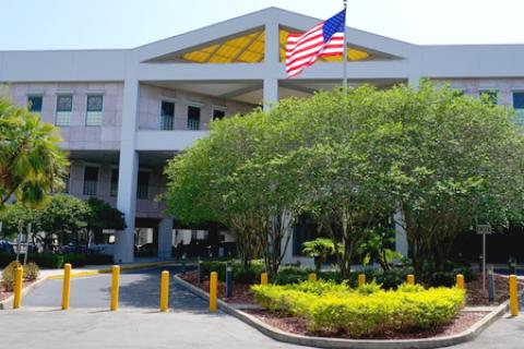 A large pink building with a U.S. flag on a flagpole, surrounded by trees and a circular driveway, under a clear sky.